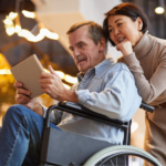 A woman stands behind a man in a wheelchair; they both are keenly focused on a tablet device.